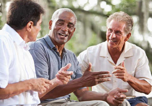 Multi-ethnic men (50s and 60s) sitting outdoors, having conversation.  Focus on African American man in middle (60s).